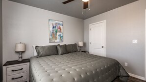 A bedroom with a tufted grey bed, a ceiling fan, a white door, and a nightstand with a lamp and drawer. - Modern Luxury Retreat | Brand-New Build | Pet-Friendly (Kingman)