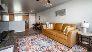 A living room with a sofa, a coffee table, and a rug. There is a ceiling fan, a dining table with chairs, and a kitchen area in the background. - Modern Luxury Retreat | Brand-New Build | Pet-Friendly (Kingman)