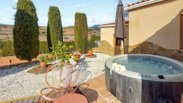 A hot tub with a stone exterior, surrounded by a patio with a chair and table, potted plants, and a view of the landscape.