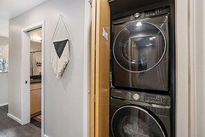 Ein Waschraum mit zwei übereinander gestapelten, frontgeladenen Waschmaschinen und Trocknern, einer Holztür und einem Gang, der zu einem Badezimmer führt.