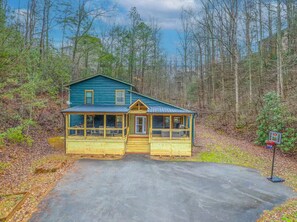 Eine zweistöckige Holzhütte mit Veranda und Basketballkorb in einem bewaldeten Gebiet.