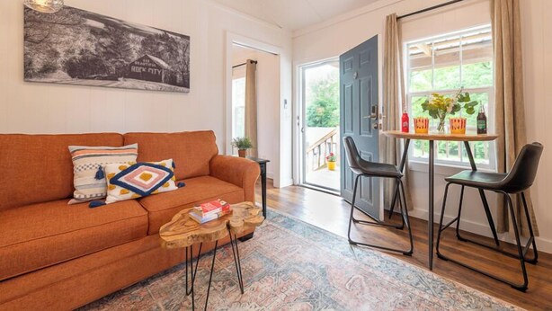 Sala de estar con sofá, mesa de centro y sillas. Se ve una puerta que da a un balcón con vistas al verdor.
