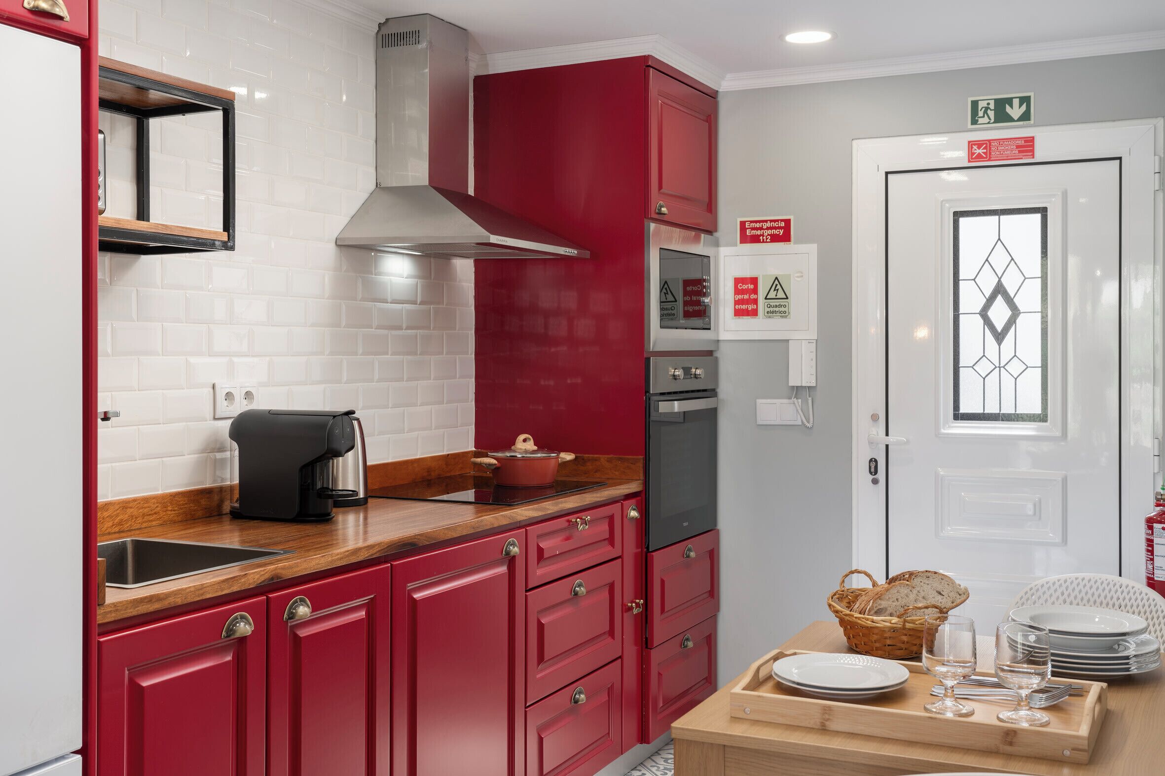 A compact kitchen with red cabinets, a stainless steel range hood, a coffee maker, and a sink. There is a door with an emergency exit sign, and a small table with plates and glasses.