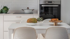 Eine moderne Küche mit weißen Schränken, einer Edelstahl-Dunstabzugshaube, einer Mikrowelle und einem Backofen. Auf der Arbeitsfläche steht eine Schale mit Obst und es sind zwei Stühle zu sehen.