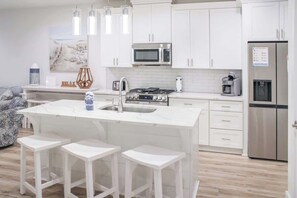 A modern kitchen with white cabinetry, a central island with stools, stainless steel appliances, and pendant lighting. - Panama City Beach 4BR | Private Heated Pool (Panama City Beach)