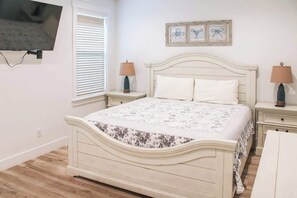 A bedroom with a bed, two bedside tables, a TV mounted on the wall, and a window with blinds. - Panama City Beach 4BR | Private Heated Pool (Panama City Beach)