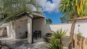 Uma área de estar coberta ao ar livre, com mesa e cadeiras, cercada por plantas e um muro de madeira.