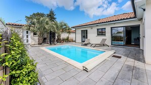 Uma piscina retangular com um deck ladrilhado, rodeada por um edifício branco com telhado ladrilhado, uma área de varanda coberta e um pequeno jardim com palmeiras.