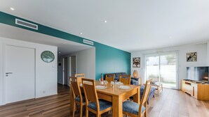 Uma área de refeições moderna com mesa e cadeiras de madeira, um relógio na parede e vista para o exterior através de uma porta de vidro.