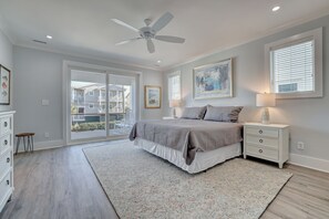 A bedroom with a bed, nightstands, a ceiling fan, a large window with blinds, and a view of a building outside. - Stunning 8BR Luxury Property Just Steps to the Beach, Pier & Restaurants! (Wrightsville Beach)