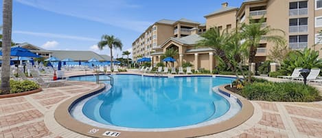 Ein Pool mit Ziegelterrassen umgeben von Liegestühlen und Sonnenschirmen, im Hintergrund ein Hotelgebäude.