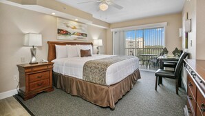 Quarto de hotel com uma cama grande, uma escrivaninha, uma cadeira, um ventilador de teto e vista para o exterior pela janela.