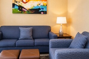 A living room with a blue sofa, matching armchair, and ottoman, a lamp on a side table, and a colorful abstract painting on the wall. - Near Disney! 1BR Deluxe Condo w/ Full Kitchen (Kissimmee)