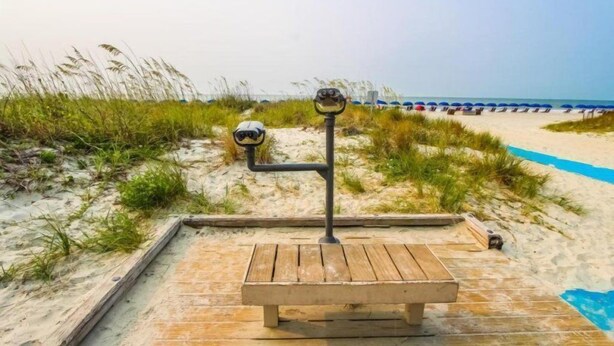 A wooden deck with a bench and binoculars on a sandy beach with dunes and beach chairs in the background. - 2BR Villa in Hilton Head | Pool Access & More! (Hilton Head Island)