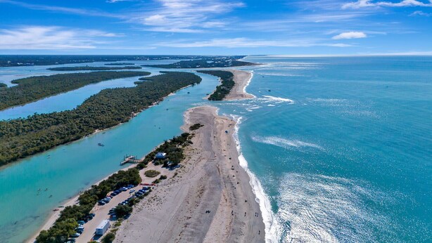 Vista aérea de una zona costera con playa, un puerto deportivo y una península boscosa.