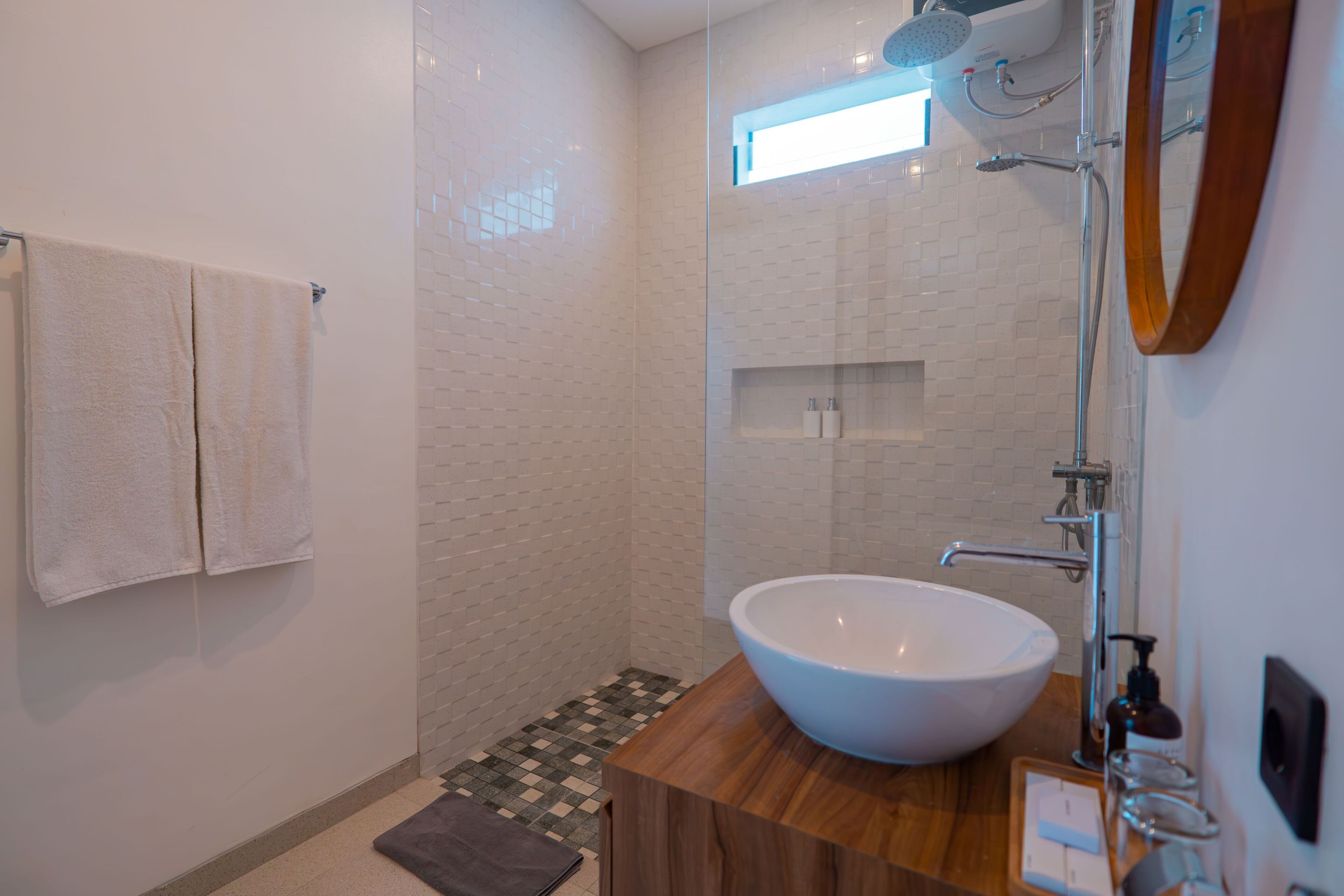 Ein Badezimmer mit einem großen weißen Waschbecken, einer Dusche und einem Spiegel. Handtücher hängen an der Wand, und ein kleines Regal beherbergt Pflegeprodukte.