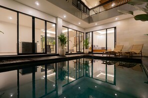 Una piscina interior moderna con paredes de vidrio, sillones de descanso y plantas en macetas.
