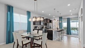 Una cocina moderna con una isla central, un comedor con mesa de vidrio y cortinas color turquesa.
