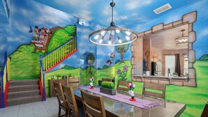 Ein Essbereich mit Wandgemälde von Mickey und Minnie Maus auf einer Brücke, einer Kristallleuchter und einer Treppe mit farbenfrohen Geländern.