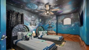 Ein Schlafzimmer im Stil von Star Wars, mit einem Bett, das ein Galaxien-Wandgemälde zeigt, einem Deckenventilator und einem Fenster mit Jalousien.