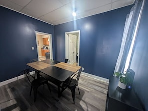 En matplats med ett bord och stolar, en krukväxt och utsikt över ett kök genom en öppen dörr.