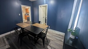 Ein Zimmer mit dunkelblauer Wand, einem Holztisch mit vier Stühlen und einer Zimmerpflanze auf einem Beistelltisch.