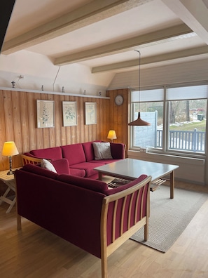 Un salon avec un canapé rouge, une table en bois, une lampe et une vue sur l’extérieur.