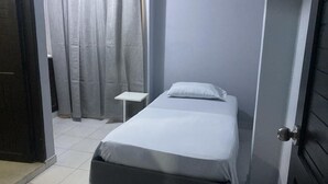 A small, single-bed room with a grey wall, a white bedspread, a white pillow, a small bedside table, and a door with a silver handle. - Hotel Tres Estrellas Fundacion (Fundación)