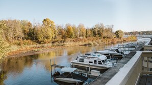 Une rangée de bateaux est amarrée dans un port de plaisance, avec une rivière et le feuillage automnal en arrière-plan.