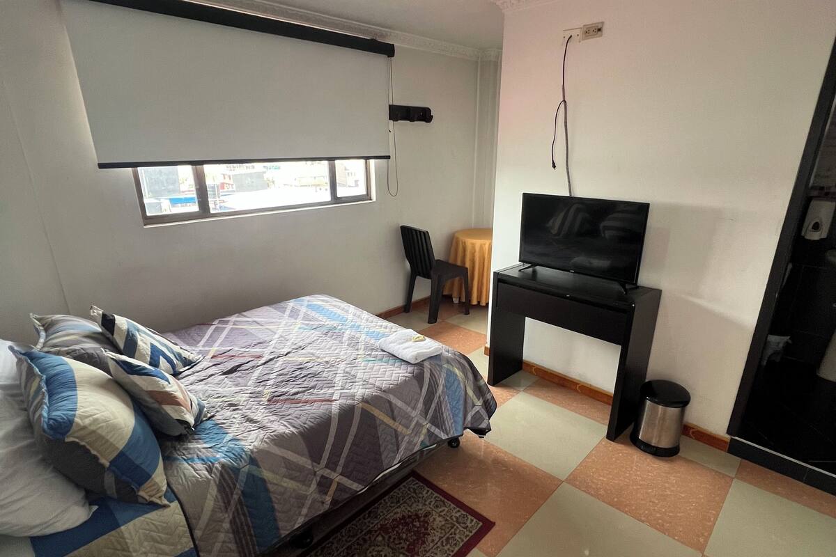 A bedroom with a bed, a TV, a small table with a chair, a window, and a ceiling light.