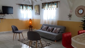 A spacious living room with a high ceiling, a grey sofa, two chairs, a red chair, a small table with a lamp, and a TV mounted on the wall. - High villa - Swimming pool - 2 min plage de Vinty - Sainte-Rose (Sainte-Rose)