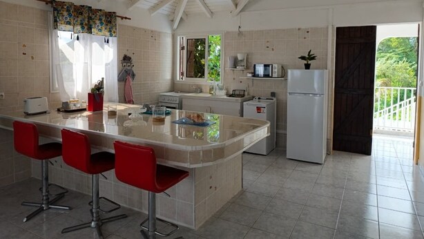 A kitchen with a central island, red bar stools, a microwave, a refrigerator, and a view of greenery outside. - High villa - Swimming pool - 2 min plage de Vinty - Sainte-Rose (Sainte-Rose)