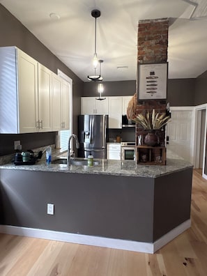 A modern kitchen with white cabinets, a central island, a brick accent wall, and a hanging light fixture. - Cozy 2-bedroom pet friendly Historic Old Cigar House (Tampa)