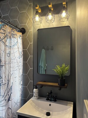 Um banheiro com parede de azulejos hexagonais, pia branca com prateleira de madeira, espelho com toalha e planta em vaso.