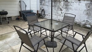 Une table de patio avec un plateau en verre, quatre chaises, un parasol et un mur de briques en arrière-plan.