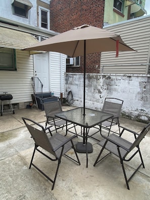 A glass-top patio table with four chairs, an umbrella, and a brick wall in the background.
