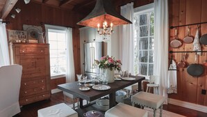 A wooden interior with a dining table set for four, a chandelier, and a kitchen area with hanging pots. - Storybook family cottage perched atop Penn Cove (coupeville)