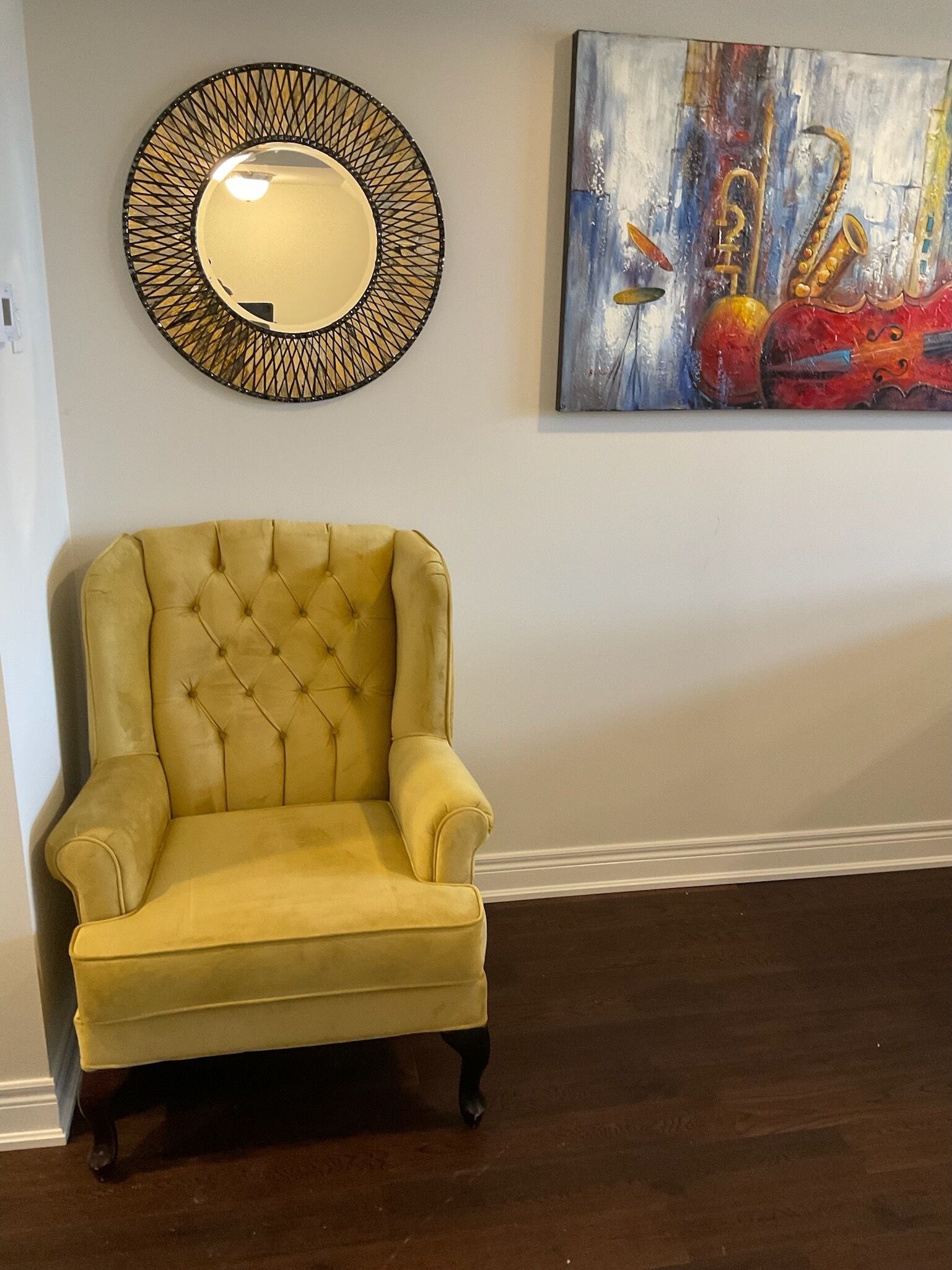 Een gele fauteuil met een gestuifte rugleuning, een decoratieve spiegel met een geometrisch frame, en een kleurrijk abstract schilderij aan de muur.