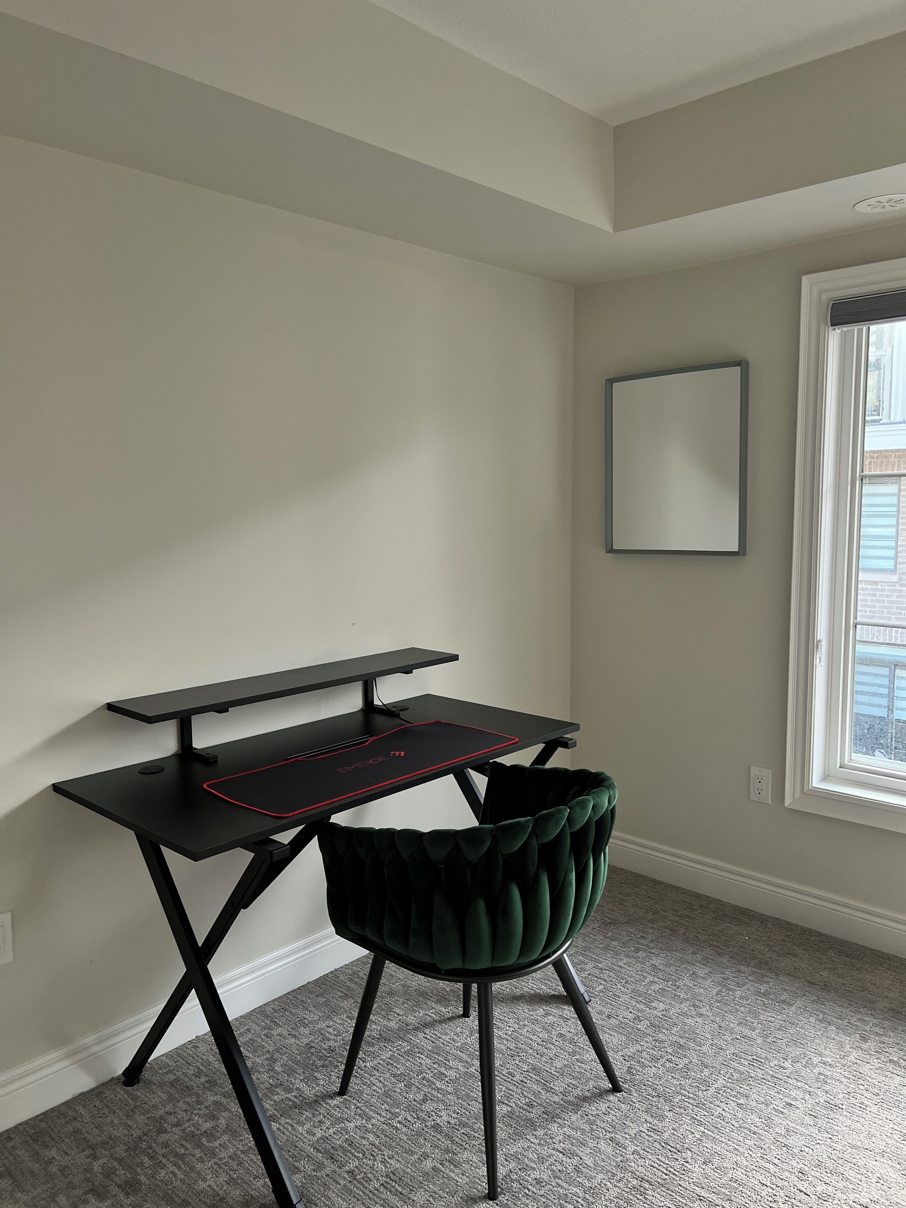 Ein Eckzimmer mit schwarzem Schreibtisch, grünem Sessel, Fenster und einem gerahmten Bild an der Wand.