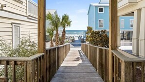 Un muelle de madera que conduce a una playa, con vista al océano, bordeado por edificios de fachadas blancas y azules.