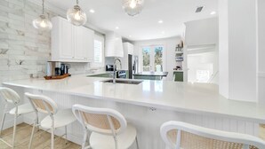 Una cocina moderna con armarios blancos, una isla grande con taburetes y iluminación colgante, además de vistas al exterior.