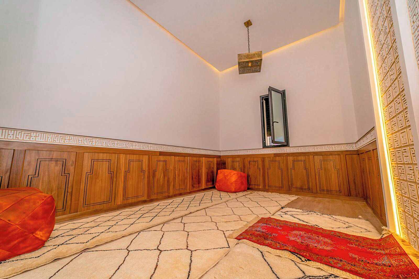 A room with a patterned floor, wooden paneling, a red ottoman, a hanging light fixture, and a red rug.