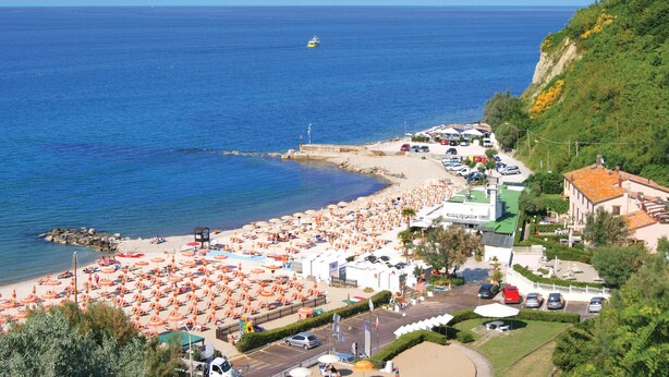 A coastal beach area with numerous umbrellas and sun loungers, a small boat in the water, and a road leading to a building with a green roof. - Lovely home in Mombaroccio (Mombaroccio)