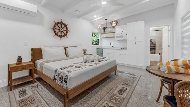Un dormitorio con cama, una mesita de noche de madera, ventilador de techo, timón de barco, una cocina con armarios y microondas, y un comedor con mesa y sillas.