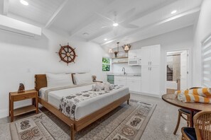 Un dormitorio con cama, una mesita de noche de madera, ventilador de techo, timón de barco, una cocina con armarios y microondas, y un comedor con mesa y sillas.