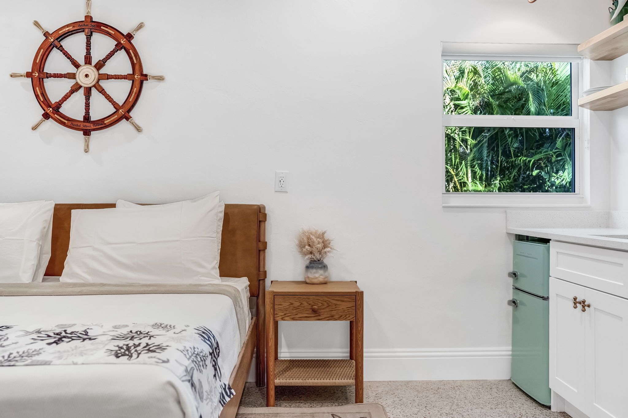 A bedroom with a bed, a nightstand with a vase, a window with a view of greenery, and a green appliance.