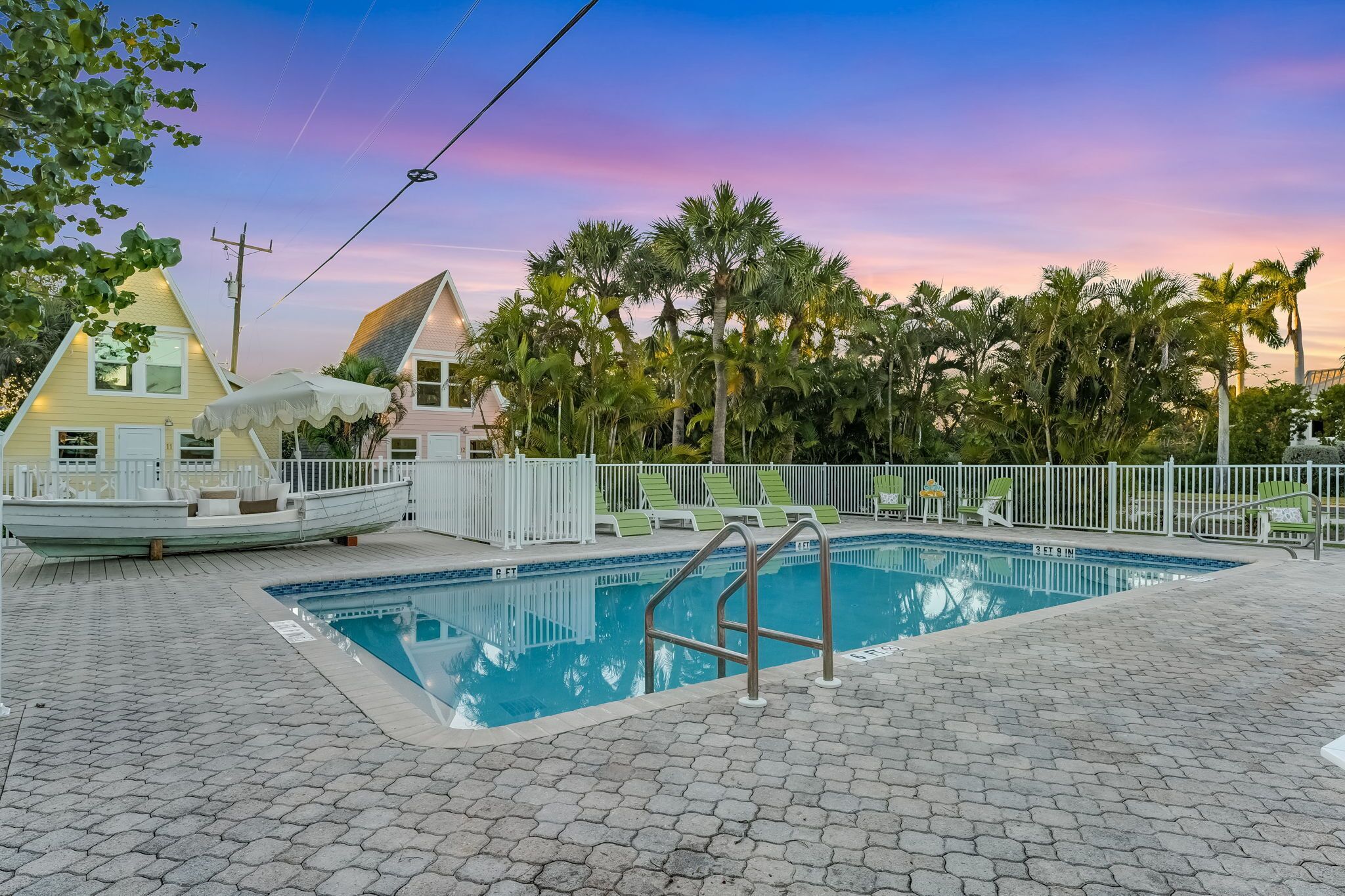 A swimming pool with a white fence, lounge chairs, and palm trees.