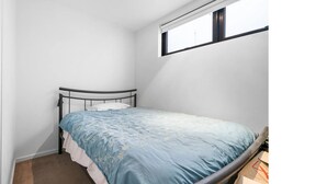Une petite chambre avec un lit simple, un cadre en métal, une couvre-lit bleue et une fenêtre avec des stores.
