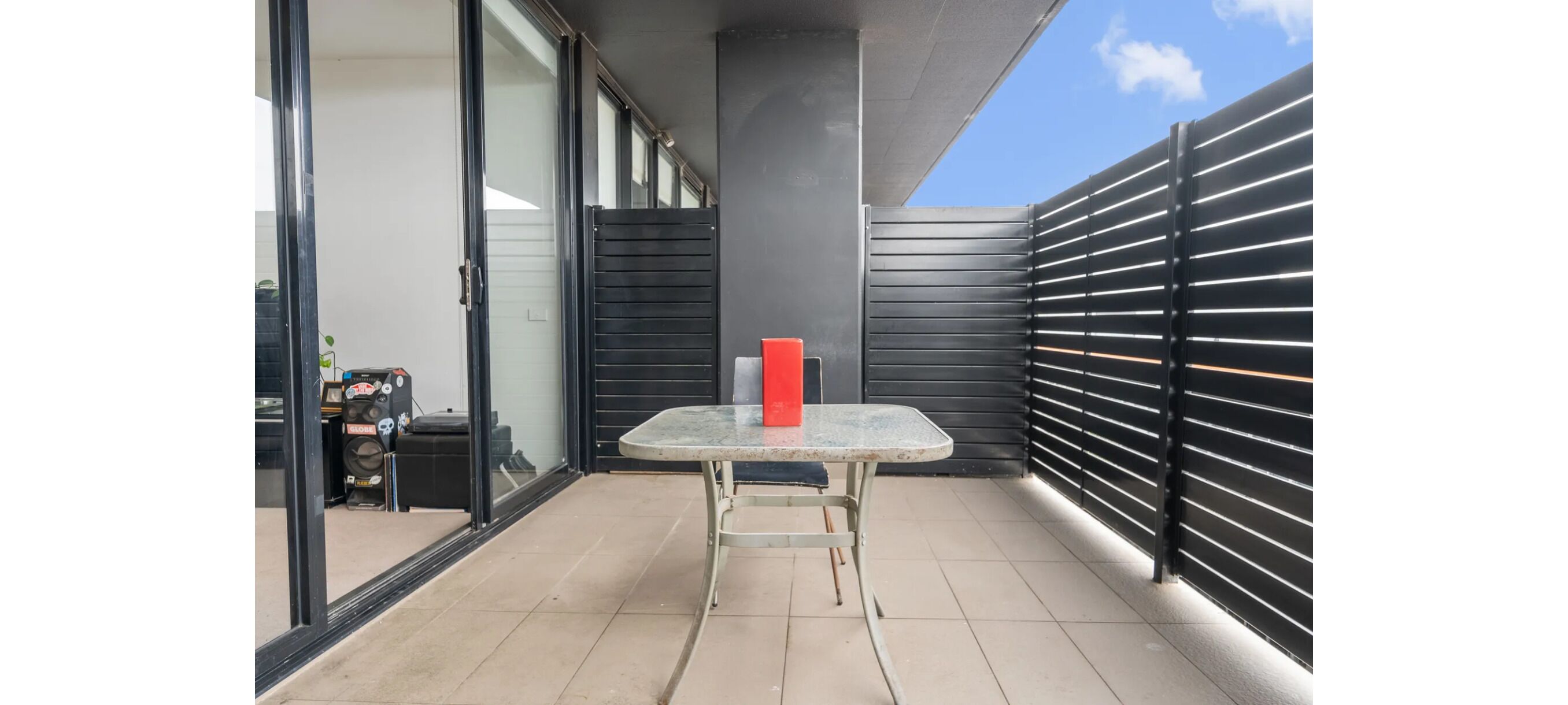 Un petit espace patio extérieur avec une table et un objet rouge posé dessus, une porte-fenêtre coulissante et une clôture en métal noir.