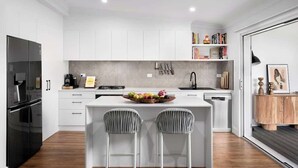 A modern kitchen with white cabinetry, a central island with bar stools, a black refrigerator, and a coffee machine. - Carrington Cottage | Walk to Cafés, Bars & Harbour (Carrington)
